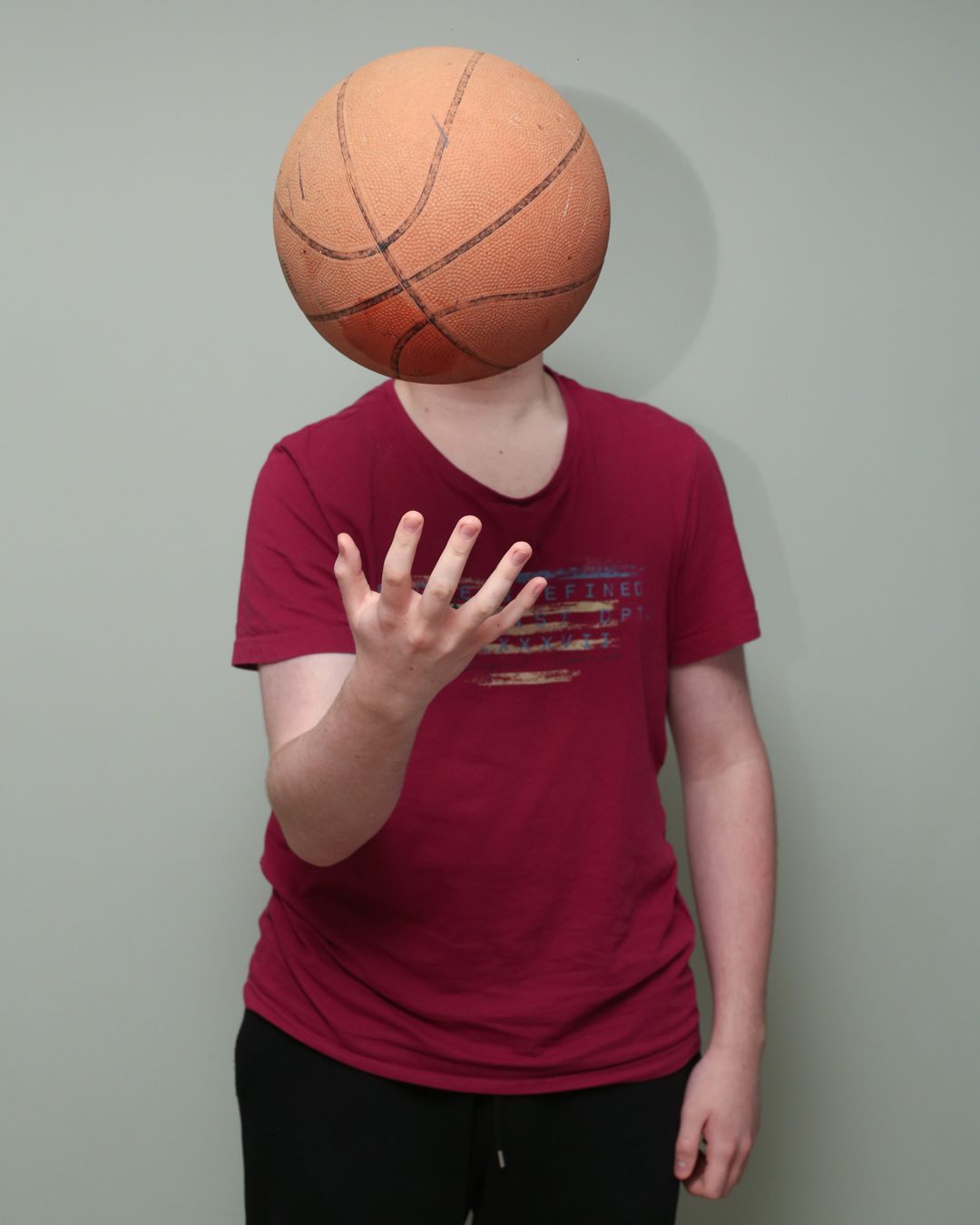 Boy in red t shirt throwing a basketball which covers his face. Portraits for shy people
