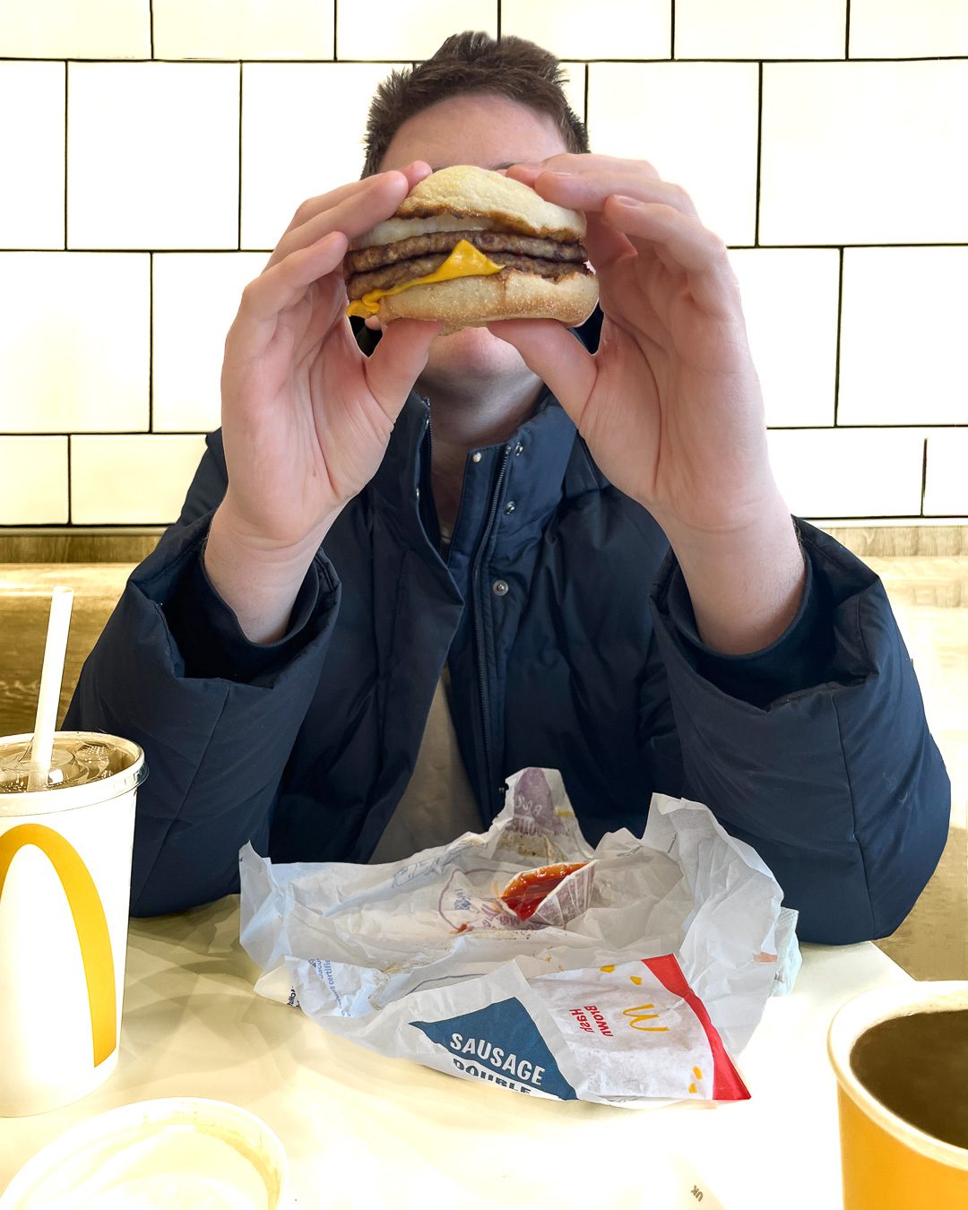 Boy eating a Macdonalds burger. White tiels in the background. His face is hidden by the burger. Portraits for shy people