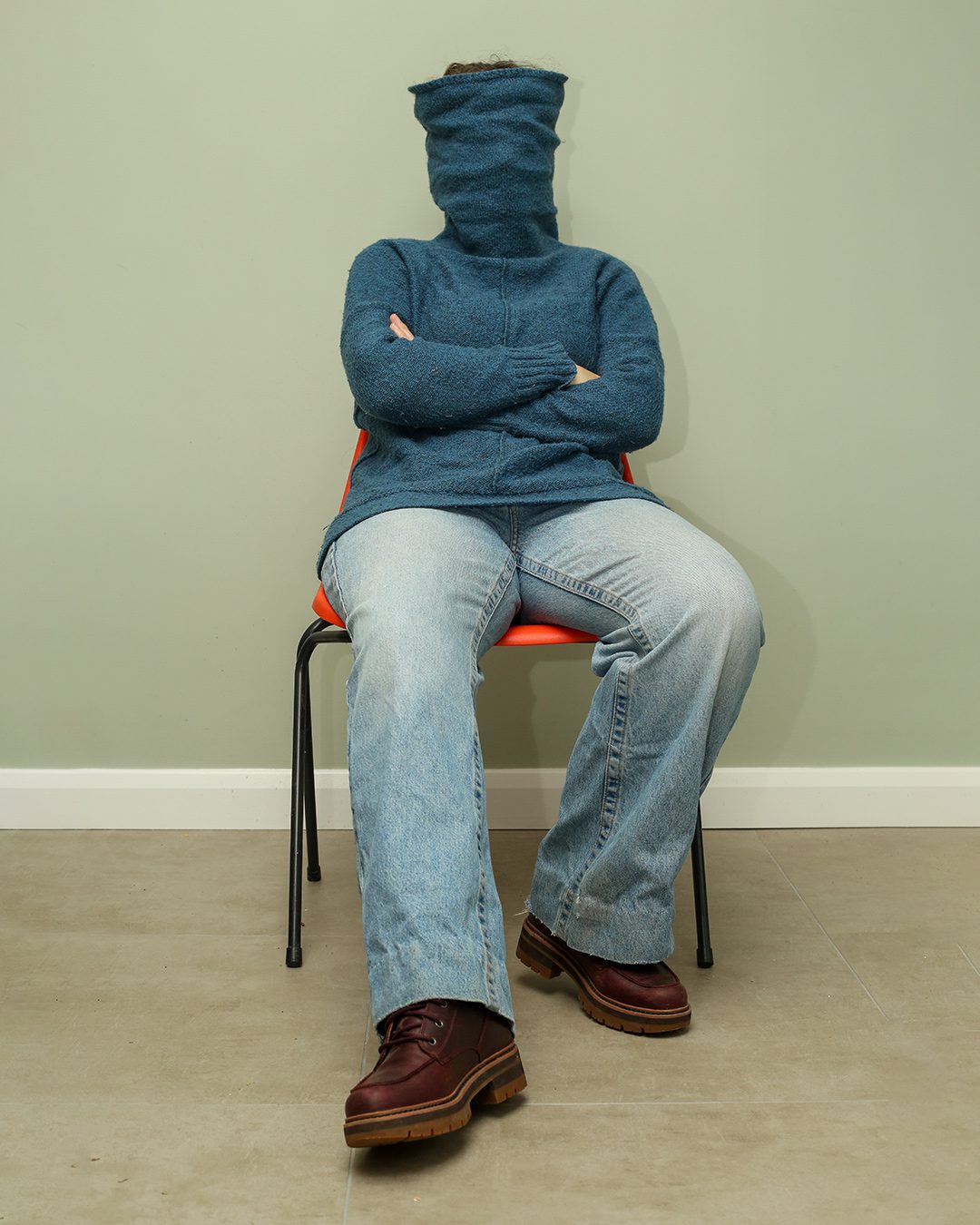 person sitting in a chair their face is obscured by a blue polo neck jumper pulled up over their face. Portraits for shy people