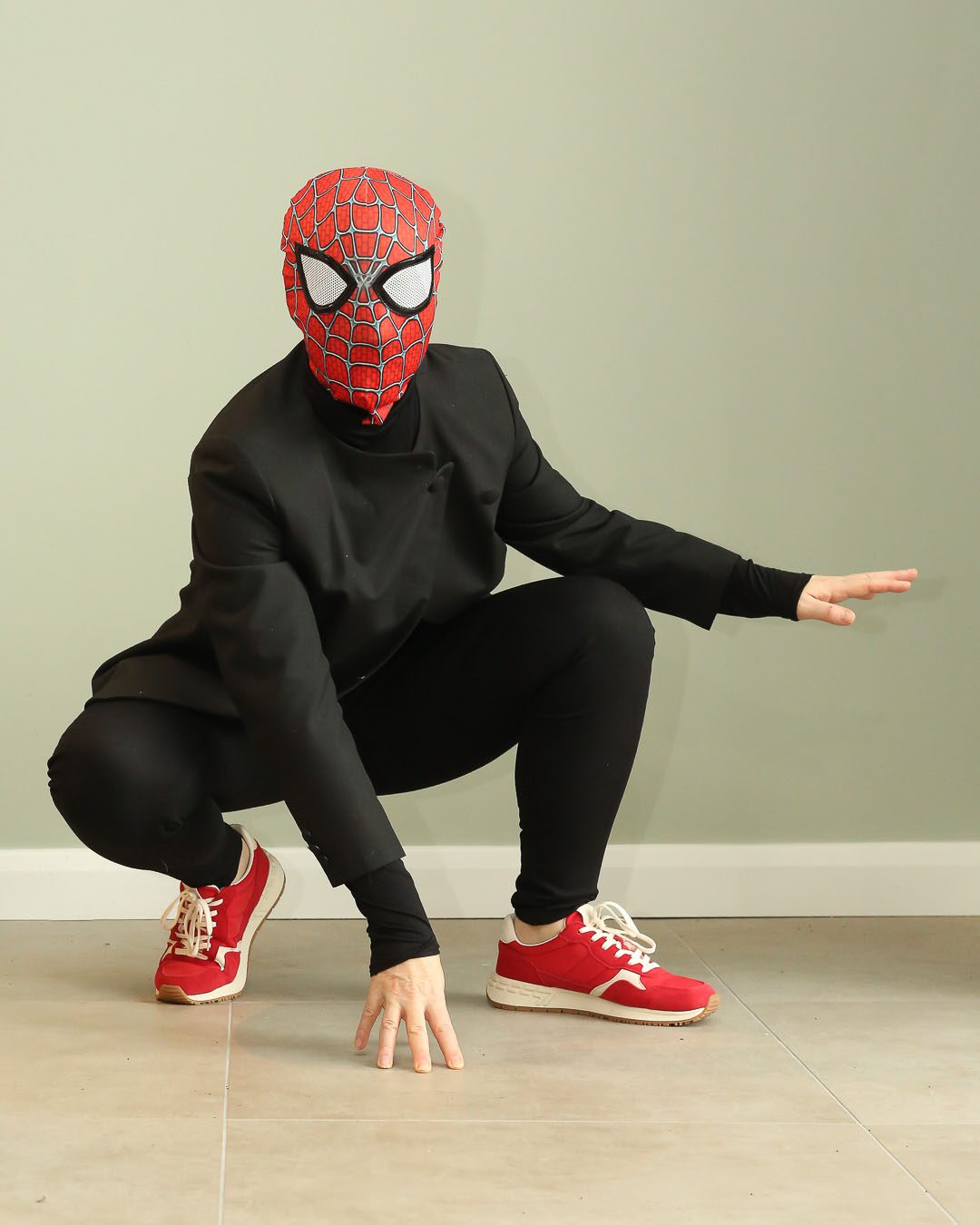 Figure in black suit with a spiderman mask in a spiderman pose against green wall. Portraits for shy people